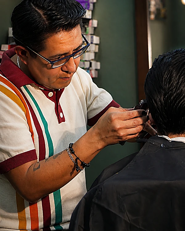 Barbero de lentes con playera de rayas de colores cortando con máquina a un cliente que está de espaldas