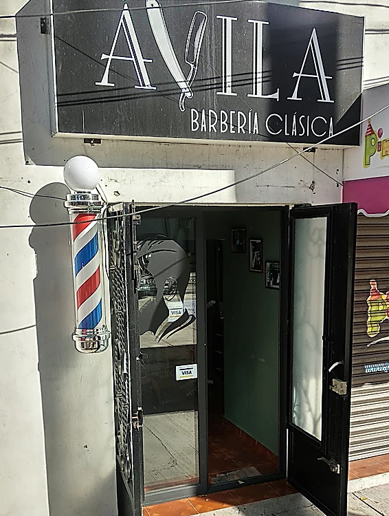 Fachada de la barbería: letrero negro con ÁVILA Barbería Clásica, poste de barbero junto a la puerta de vidrio abierta