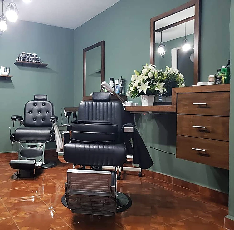 Interior de la barbería: dos sillones clásicos negros, uno capitoné, espejos con marco de madera, gabinete de cajones, flores blancas y piso de mosaico terracota