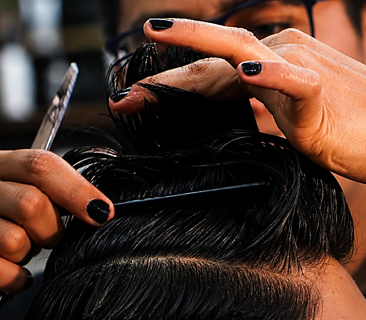 Manos del barbero con uñas negras sosteniendo tijera y peine sobre el cabello oscuro de un cliente