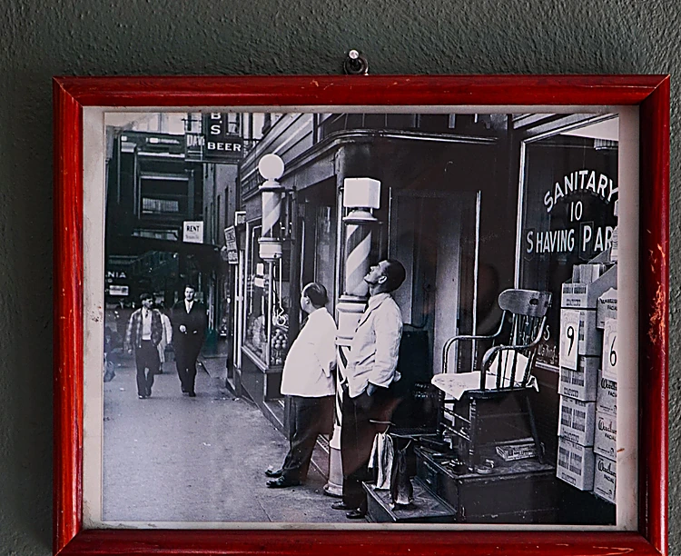 Fotografía antigua en blanco y negro de una barbería con el letrero Sanitary Shaving Parlor, en un marco rojo colgado del muro