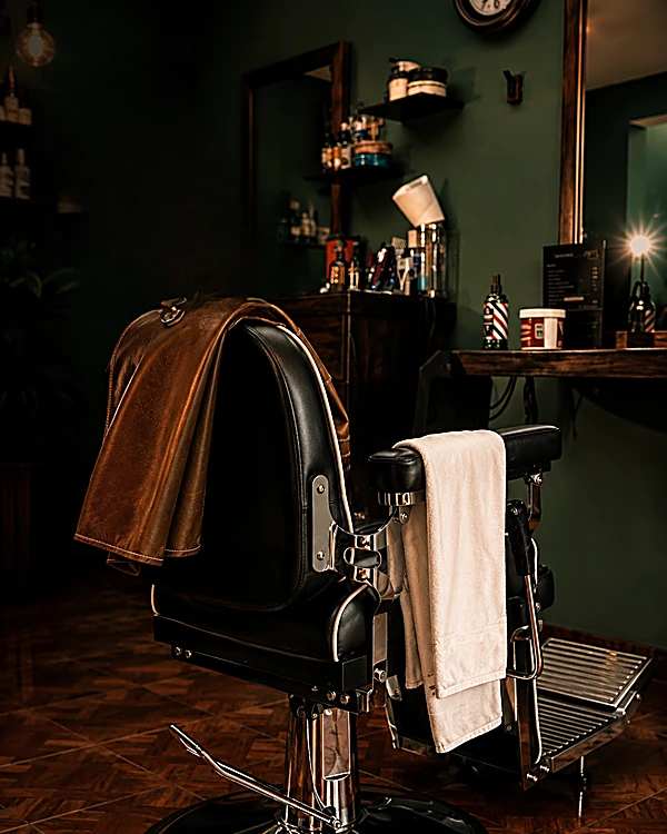 Sillón de barbero negro con una toalla blanca y una chamarra de piel café en el respaldo, frente al muro verde botella y la repisa con productos